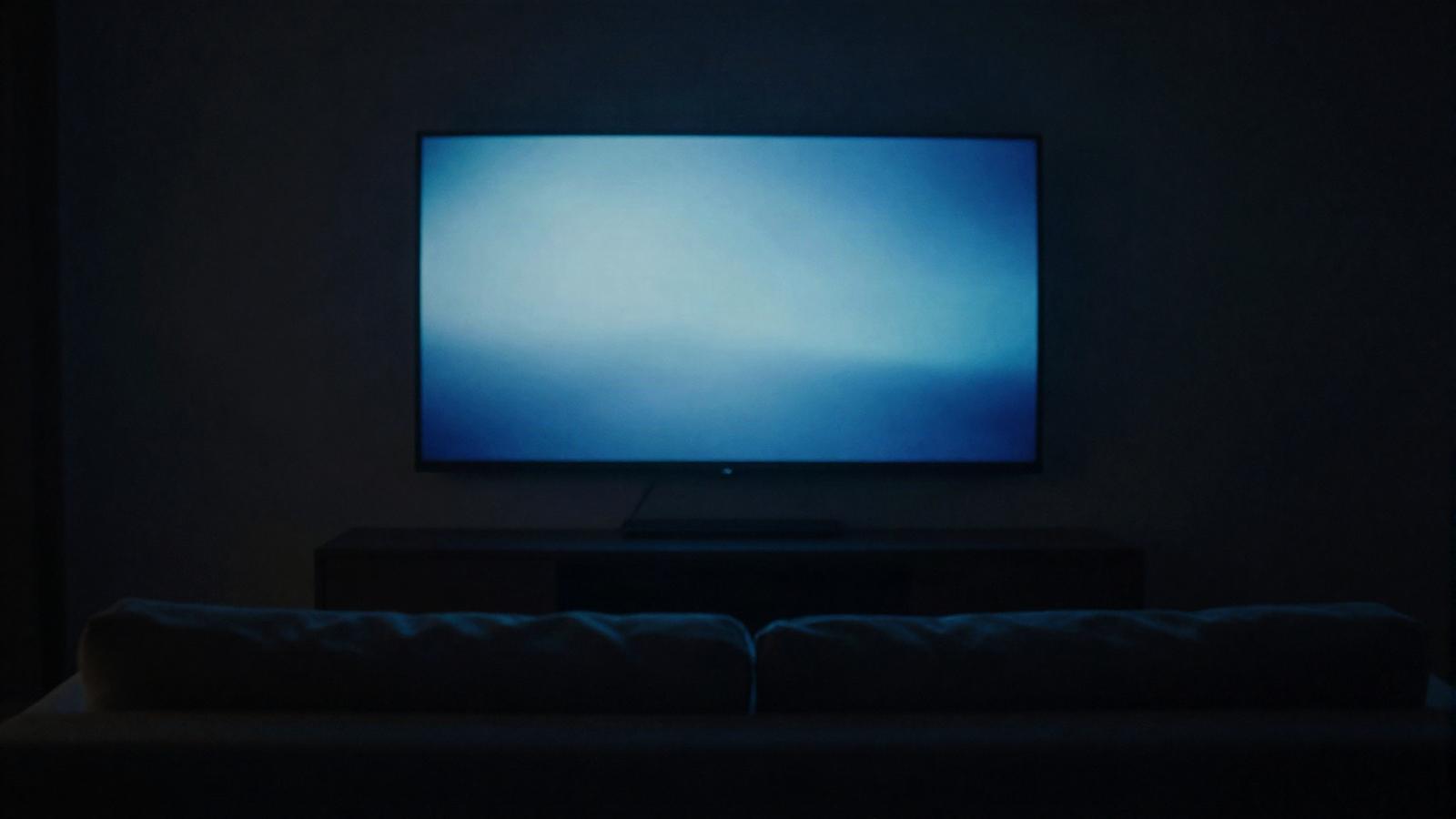 A large wall mounted television glowing in a dark living room, seen from behind a sofa